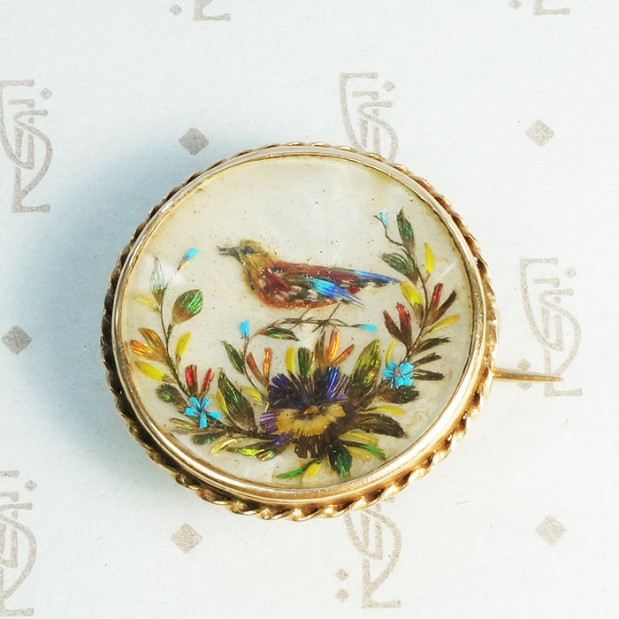 a brooch holding a scene of a bird in flowers made out of colorful feathers on a mother of pearl background in a 14k yellow gold frame